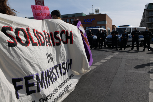Eine Gruppe von Menschen marschiert auf einer Straße, hält ein Banner mit der Aufschrift "Solidarität und Feminismus" und hat im Hintergrund parkende Fahrzeuge, Gebäude, eine Schüsselantenne und einen klaren blauen Himmel.