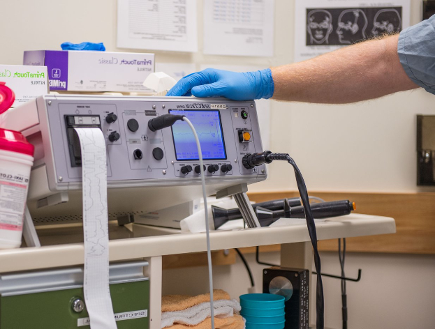 Eine Person in blauen Handschuhen arbeitet an einem medizinischen Gerät auf einem Tisch, der mit verschiedenen Gegenständen übersät ist, mit Papieren an der Wand in einer Krankenhausumgebung.