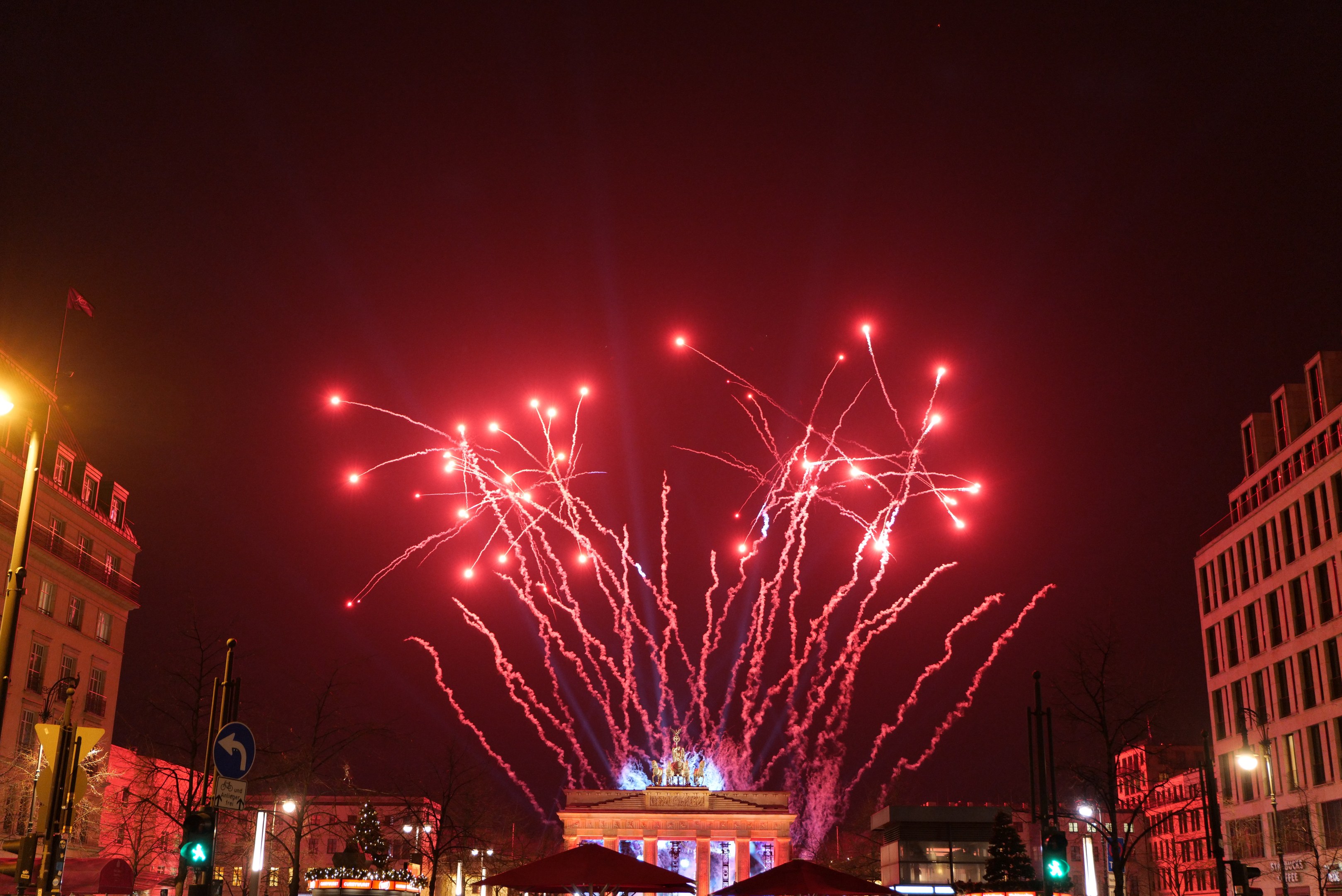 Eine belebte Stadtstraße an einem Neujahrsabend in Berlin mit Gebäuden, Bäumen, Laternenpfählen, Verkehrsampeln, Schildern, Zelten, Menschen und einem prächtigen Feuerwerk am Himmel.