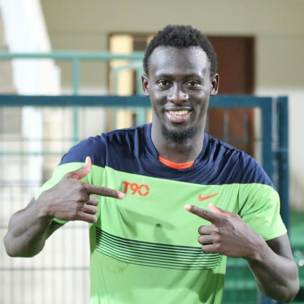 Ein Mann in einem grünen und blauen Hemd, der in die Kamera lächelt und zeigt, mit einem unscharfen Geländer, Wand und Tür im Hintergrund.