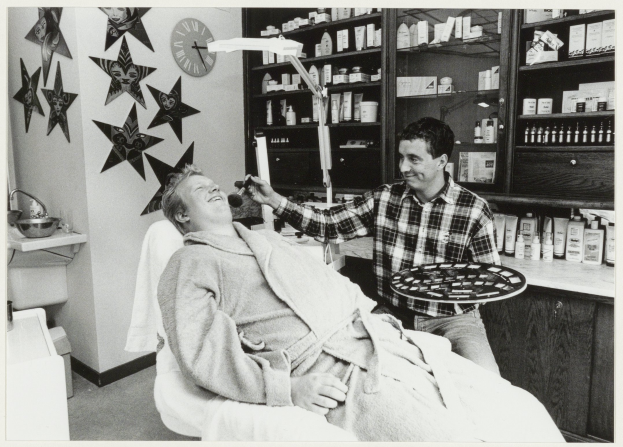 Ein Mann lächelt in einem Zahnarztstuhl, während ein anderer Mann neben ihm steht und ein Tablett hält, mit einem Waschbecken, Uhr und Regalen mit Flaschen im Hintergrund.