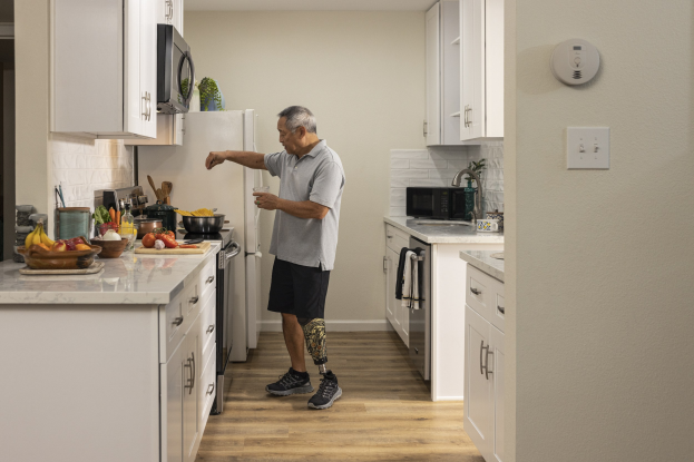Ein Mann steht in einer Küche mit wei├čen Schr├Ąnken, h├Ąlt ein Glas in der Hand, w├Ąhrend verschiedene Küchenutensilien, darunter Obst und Gemüse, auf einer Arbeitsplatte zu seiner Linken sind.