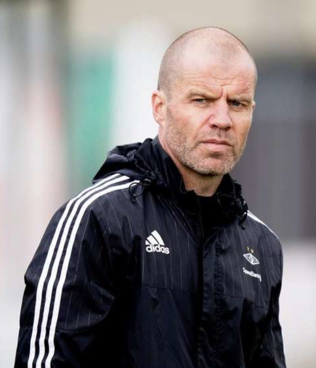 Baldiger Trainer in marineblauer Jacke mit weißen Streifen und weißem Kragen auf einem Fußballfeld stehend, Hände verschränkt und ernster Gesichtsausdruck.