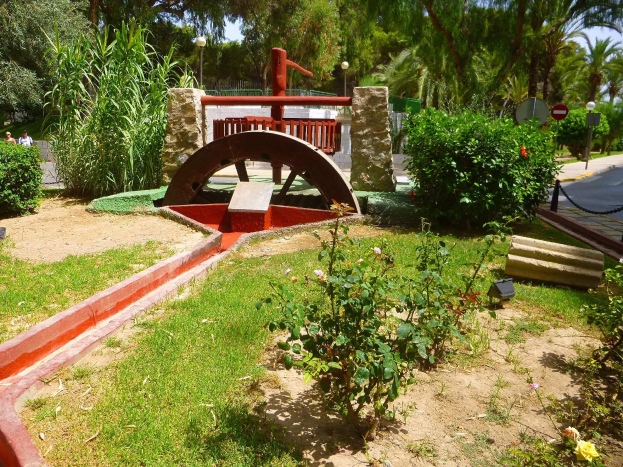 Ein Minigolf-Platz in einem Park, umgeben von Grün, mit einer Straße und Fußgängern auf der rechten Seite und Bäumen im Hintergrund bei klarem Himmel.