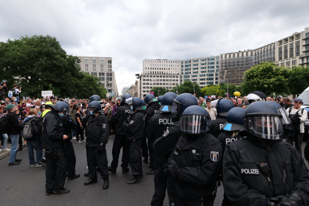 Eine große Gruppe von Polizisten steht vor einer Menschenmenge auf einer Straße, die von Bäumen und Gebäuden gesäumt ist, unter einem bewölkten Himmel während einer Demonstration in Berlin, Deutschland. Einige in der Menge halten Kameras.