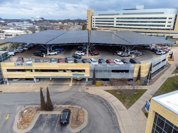 Luftaufnahme eines solarpanelbedeckten Parkplatzes mit parkenden Autos, umgeben von Gebäuden, Bäumen, Grüsser, Straße und Hühlen unter einem bewölktem Himmel.