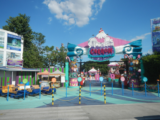 Eine Karussellfahrt am Parkeingang, mit einer Straße darunter, einem Zaun, Buden, Schildern, Bäumen und einem bewölkten Himmel im Hintergrund.